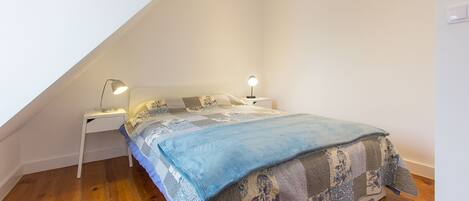 A bedroom with a sloped ceiling, a bed with patterned bedding, two bedside lamps, and wooden flooring.