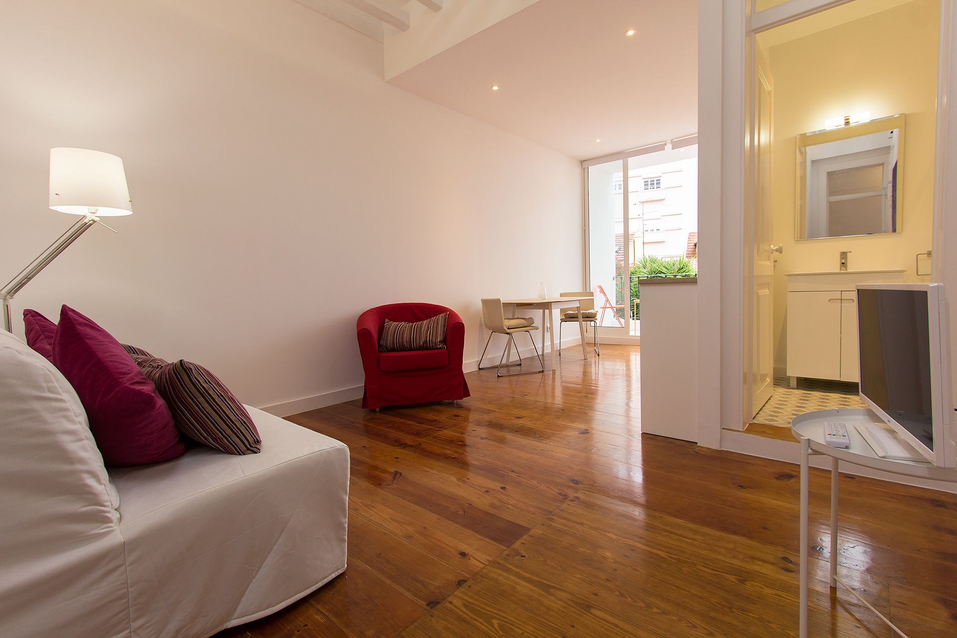 Ein modernes Wohnzimmer mit einem roten Sessel, einem weißen Sofa mit Kissen, Holzboden und Blick ins Freie durch eine Glastür.