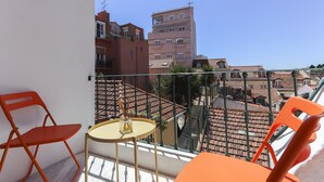 Un balcón con dos sillas anaranjadas y una pequeña mesa redonda, con vista a un paisaje urbano de edificios con tejados de teja.
