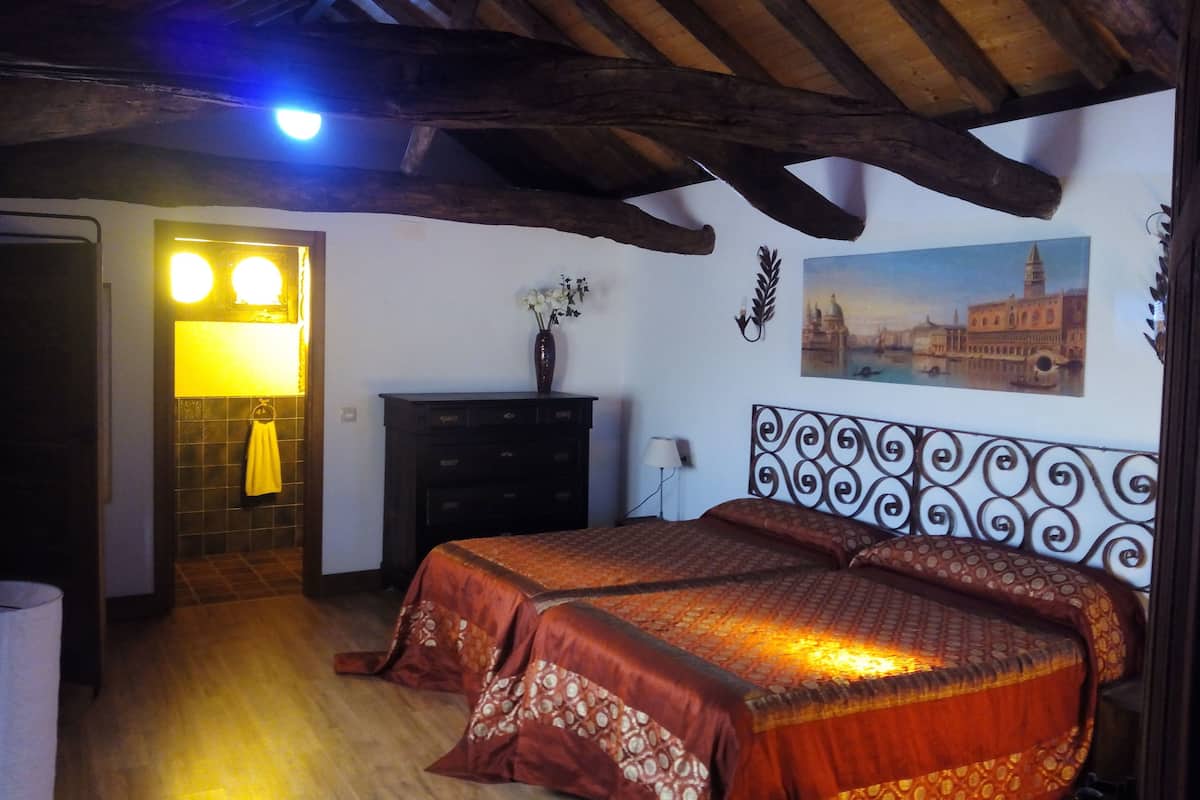 Un dormitorio con techo de madera, una cama con ropa de cama estampada, un aparador, un florero con flores y un cuadro de un paisaje urbano.