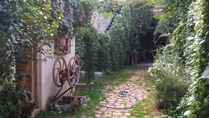 Un sendero pavimentado atraviesa un jardín con una escultura metálica rústica, rodeado de abundante vegetación y un edificio con tejado de azulejos.