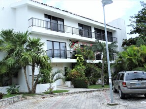 Un edificio de dos pisos con balcones, rodeado de exuberante vegetación y un automóvil estacionado.