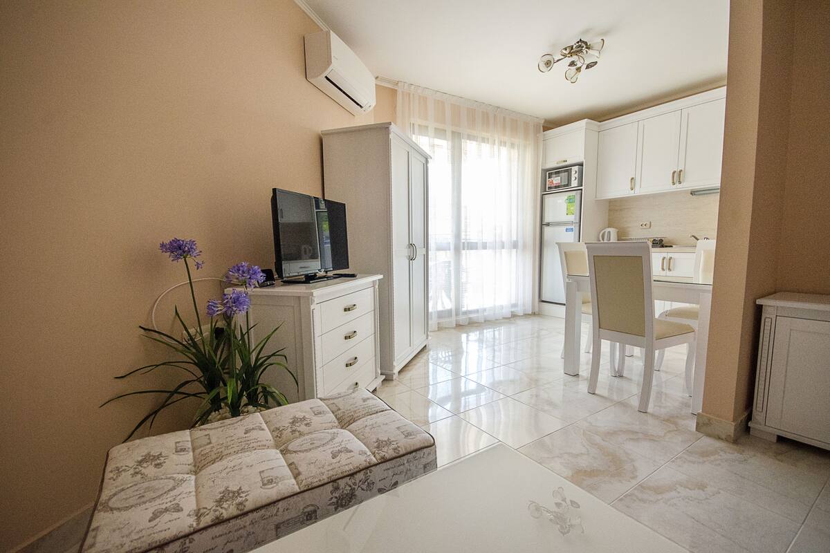 A modern kitchen with white cabinets, a dining area with a white table and chairs, and a living area with a sofa and a television.