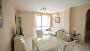 A dining area with a glass table, white chairs, a sofa, and a small table with a plant. - Apartments in Complex Poseidon (Nessebar)