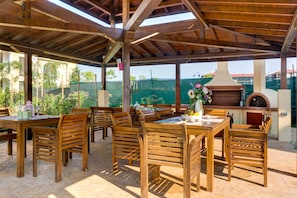 An outdoor patio with wooden tables and chairs, a brick oven, and a view of a green fence and buildings in the background. - Apartments in Complex Poseidon (Nessebar)