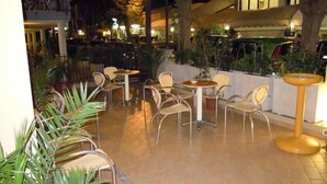 Un área de patio al aire libre con varias mesas y sillas redondas, rodeada de plantas en macetas y árboles.