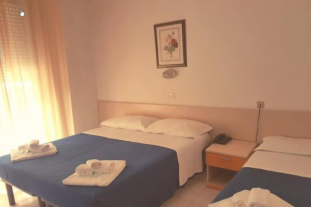 A hotel room with two beds, each with a blue bedspread and white pillows. There is a framed picture on the wall above the beds, a bedside table with a phone, and towels neatly folded on each bed.