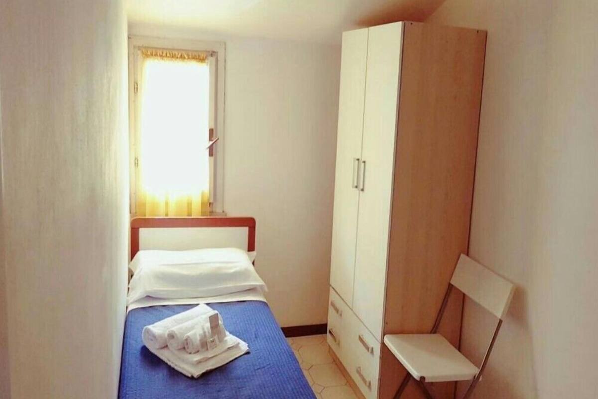 A small, single-bed room with a wooden wardrobe, a chair, and a window with curtains.