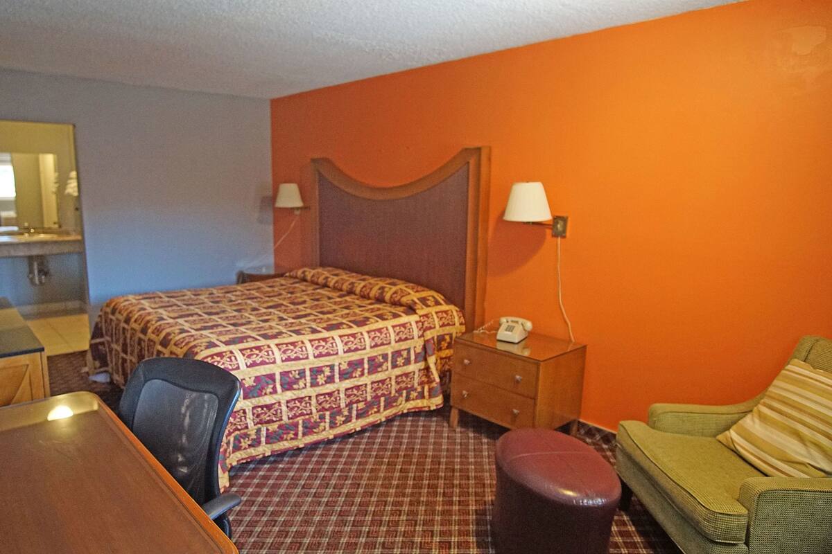 A hotel room with a bed, a desk, a chair, a small table, a nightstand, a lamp, and a couch.