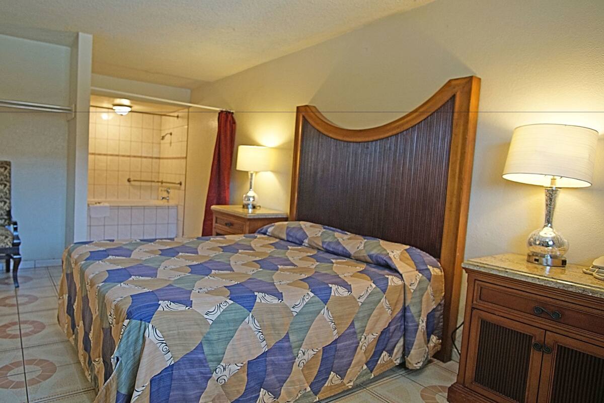 A hotel room with a large bed, a chair, a nightstand, a lamp, and a bathroom visible through an open door.