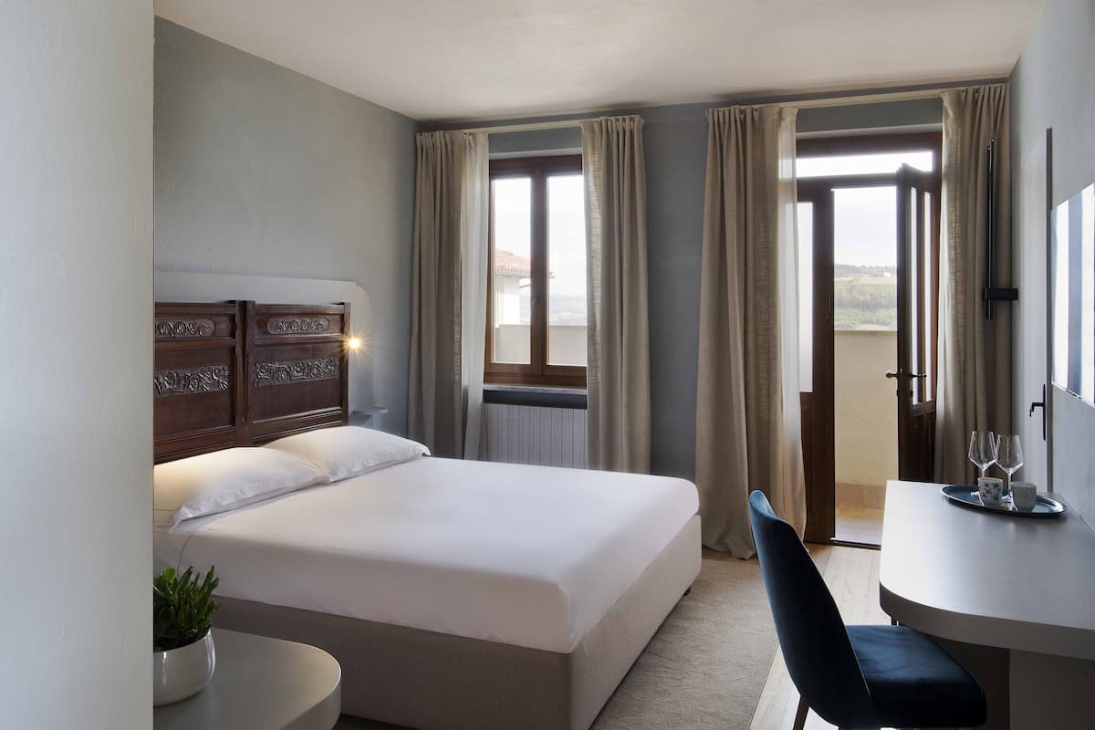 A hotel room with a bed, a desk with a tray of glasses, and a view of the outdoors through the window.