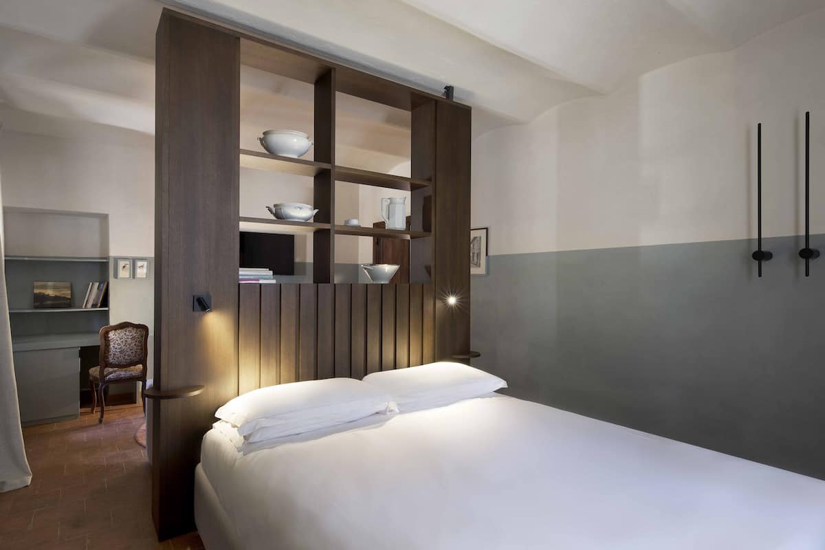 A hotel room with a bed, a built-in shelf unit with kitchenware, a chair, and a desk.