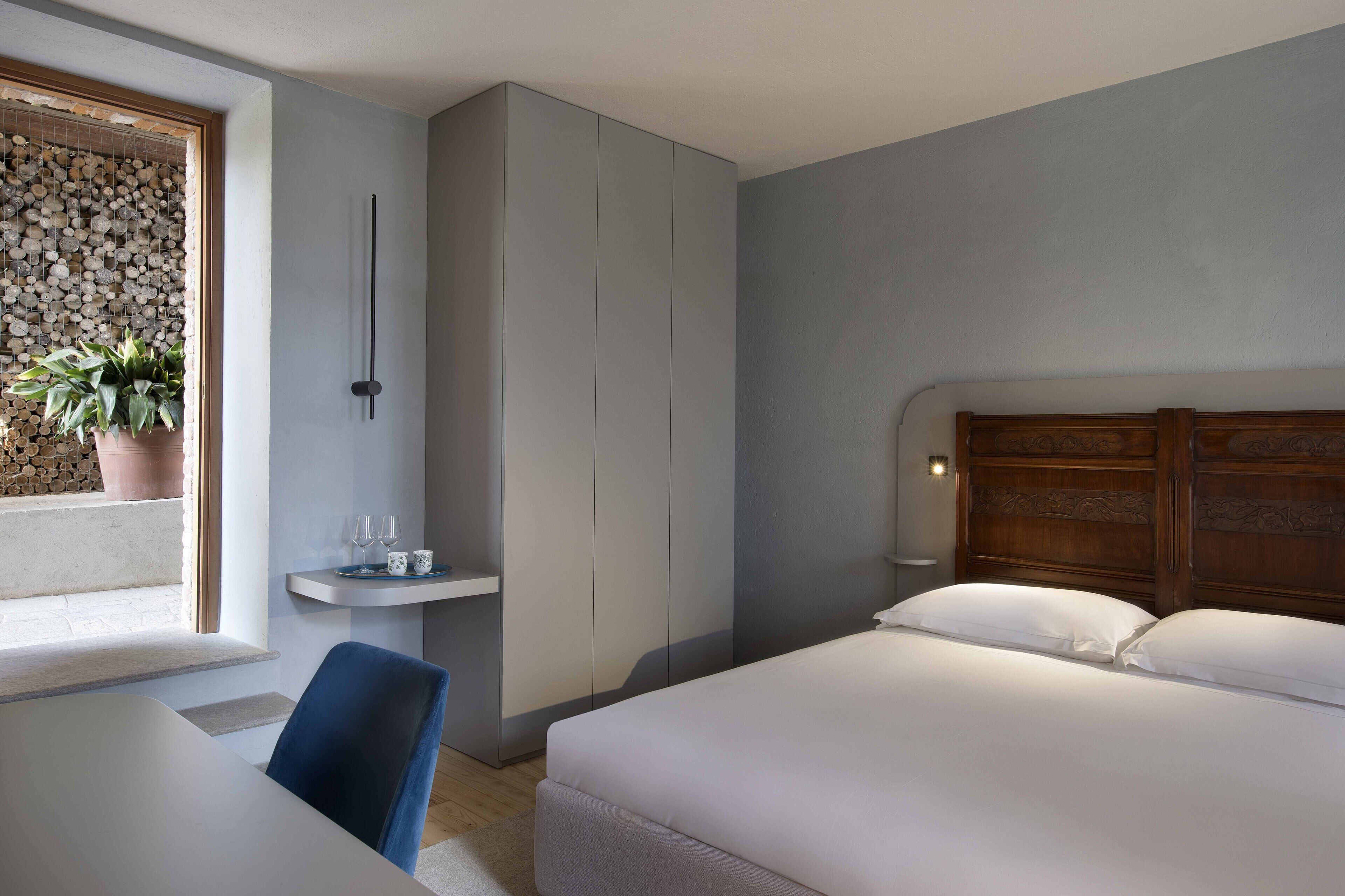 Habitación de hotel moderna con cama, escritorio, silla, mesita con vasos, planta en maceta y lámpara de pared.