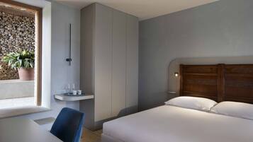 Habitación de hotel moderna con cama, escritorio, silla, mesita con vasos, planta en maceta y lámpara de pared.
