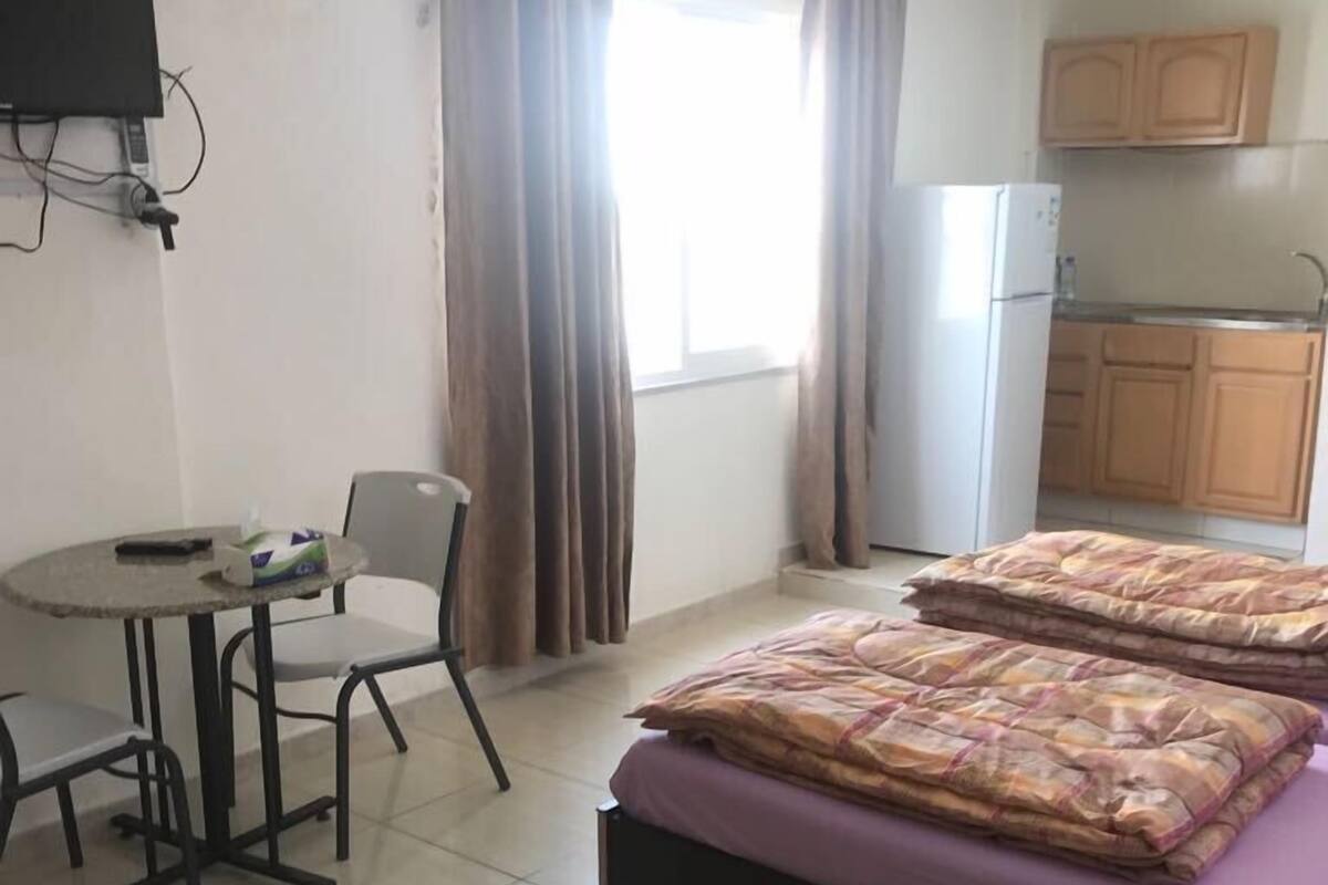 Una habitación con dos camas, una mesita con sillas, una kitchenette y un ventanal con cortinas.
