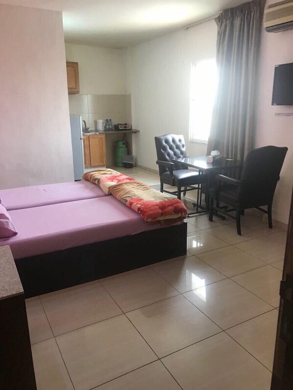 A compact room with a bed, a small desk, and a chair. There is a kitchenette in the background.