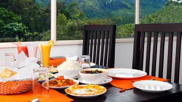 A dining table with various dishes and drinks, set against a backdrop of a mountainous landscape through large windows. - Pine Tree Munnar (Devikolam)