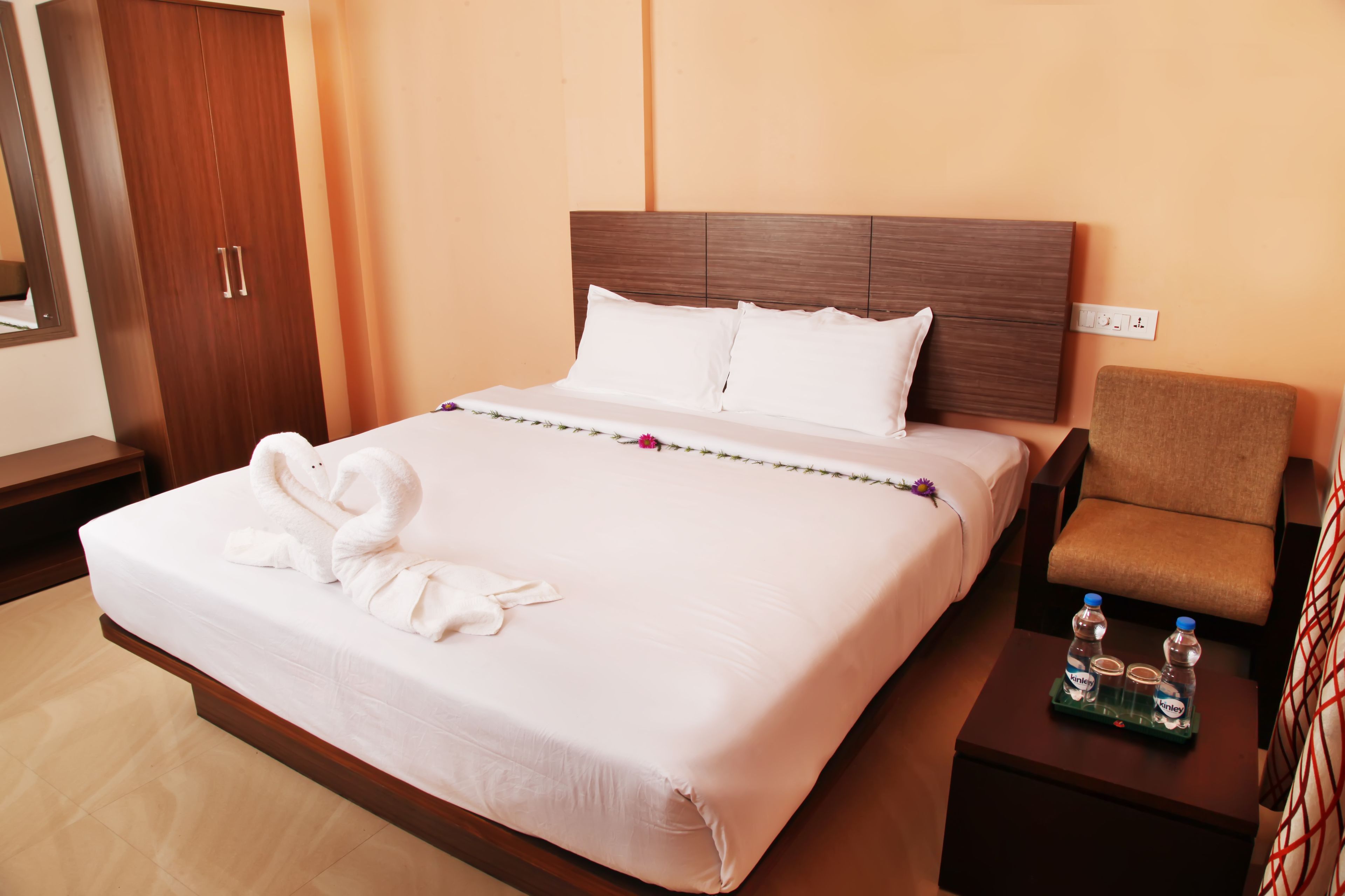 Habitación de hotel con una cama grande, un armario de madera, un sillón pequeño y una mesita de noche con botellas de agua.