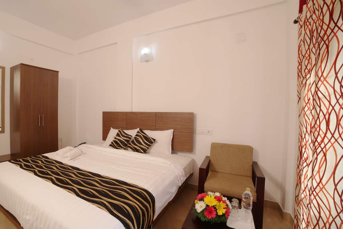 A hotel room with a bed, a wooden wardrobe, a chair, a small table with a flower arrangement, and a mirror.