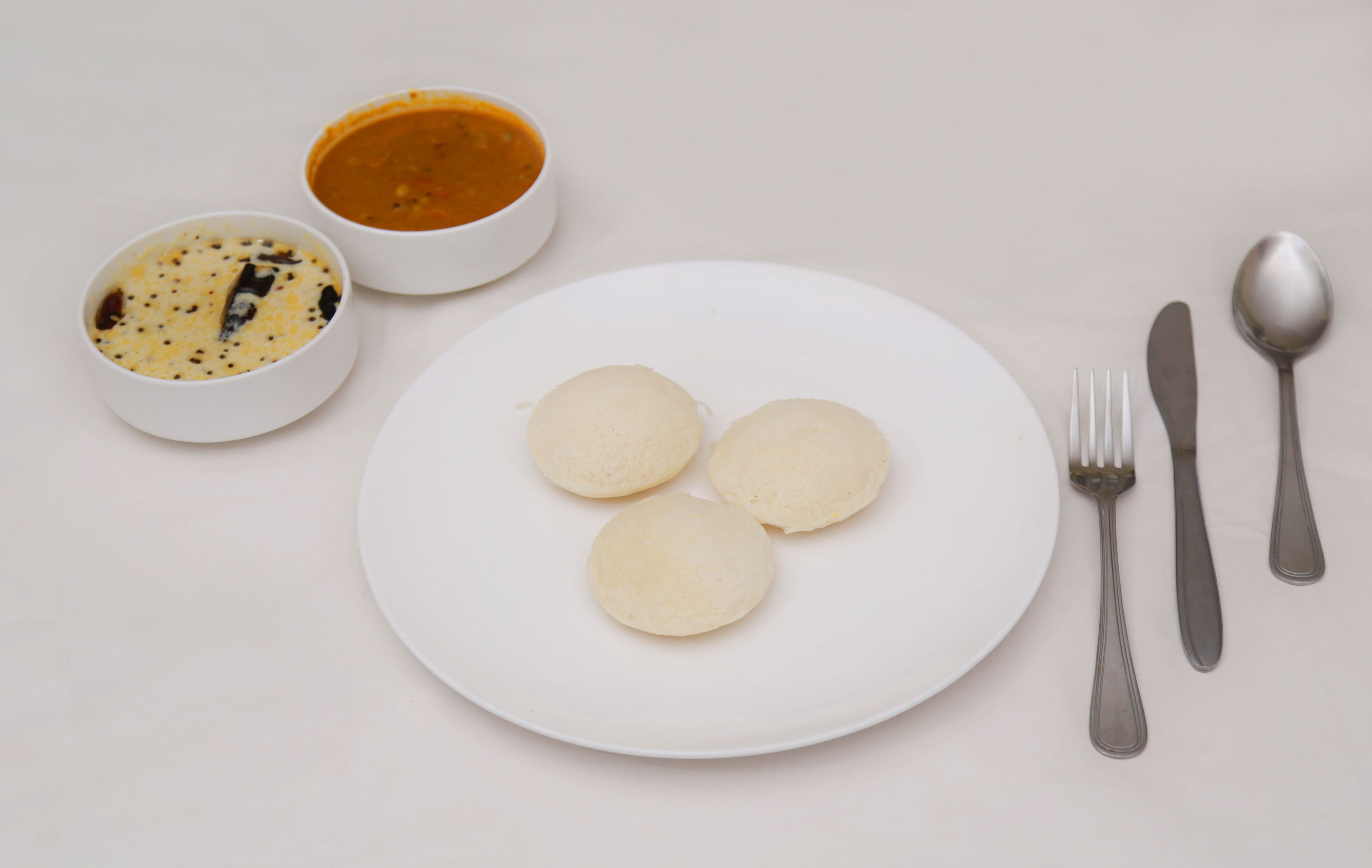 Un plato blanco con tres porciones redondas, un tenedor, un cuchillo, una cuchara y dos tazones con comida.