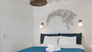 A double bed with a blue quilt, two wall-mounted lights, a decorative wall piece, and a hanging light fixture. - Nikos rooms (Mykonos)