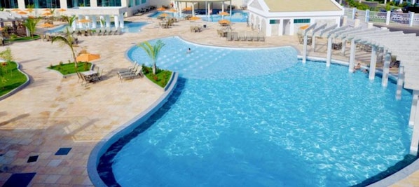 A large outdoor swimming pool with lounge chairs and umbrellas, surrounded by a landscaped area with palm trees.