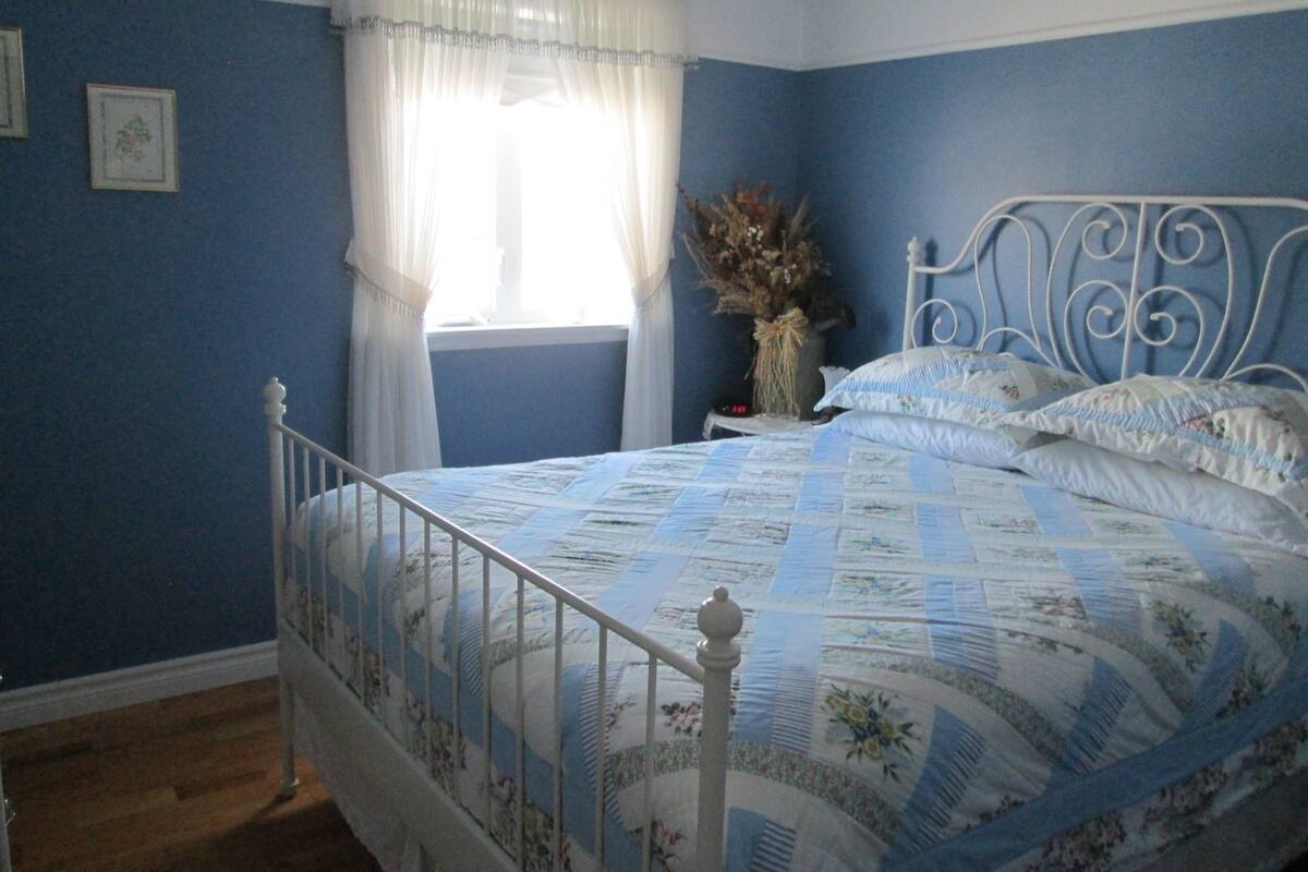 Un dormitorio con cama, mesita de noche, ventana con cortinas y un jarrón con flores secas.