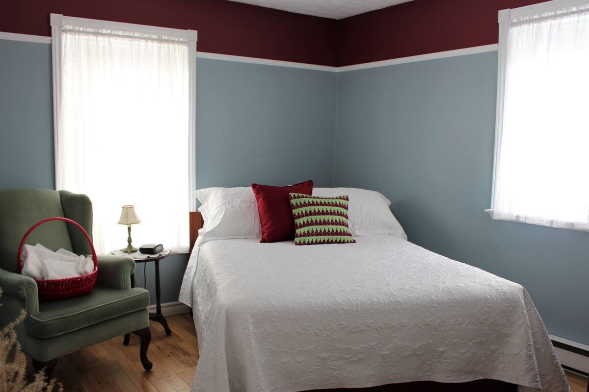 Un dormitorio con una cama, un sillón verde, una mesita de noche con una lámpara y una cesta.