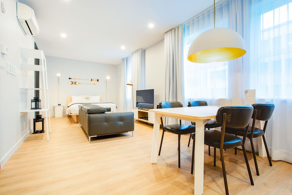 A modern hotel room with a dining area, a sofa, a TV, and a large window with curtains.