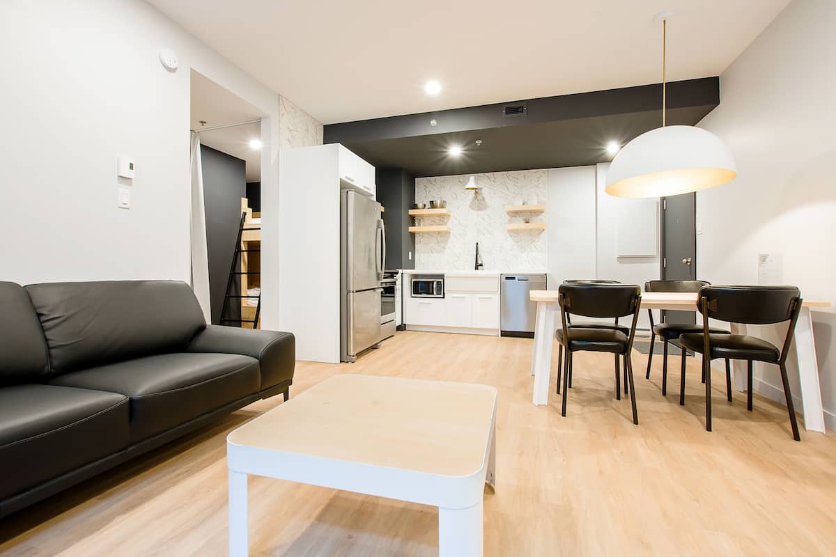 A modern living room with a sofa, a dining table with chairs, a kitchen area with a refrigerator, microwave, and shelves.
