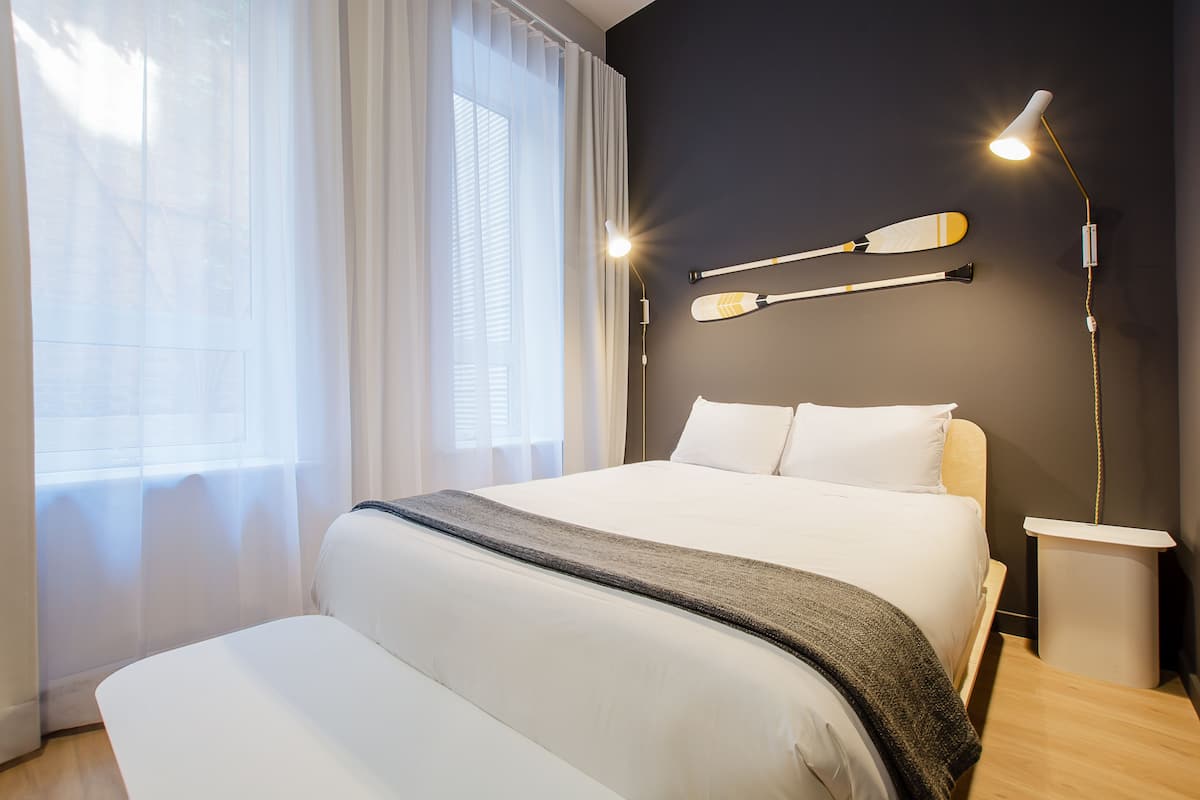 A modern hotel room with a large bed, a bedside table, a lamp, and a wall-mounted paddleboard.