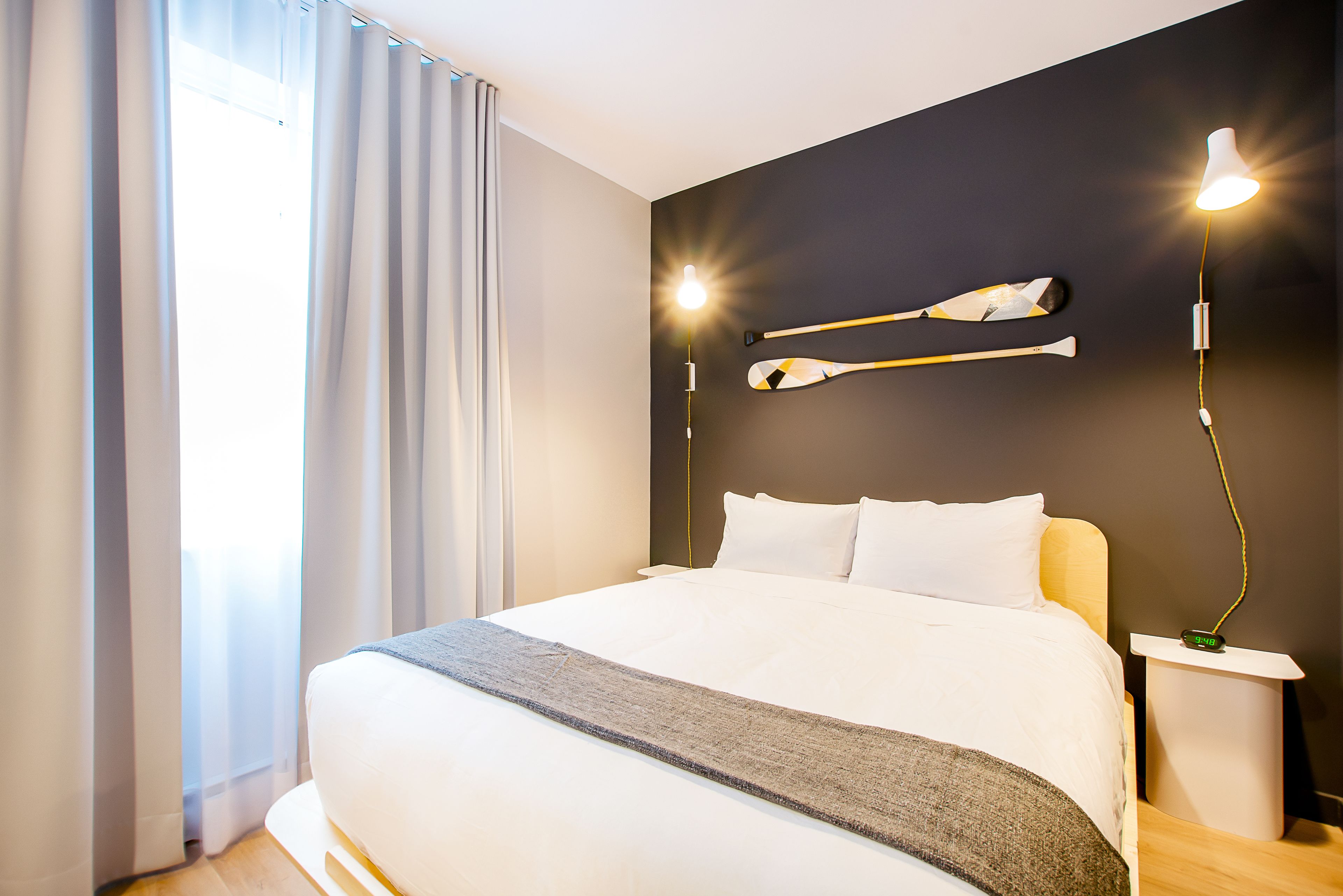 A modern hotel room with a large bed, a bedside table with a lamp, and a wall-mounted light fixture.