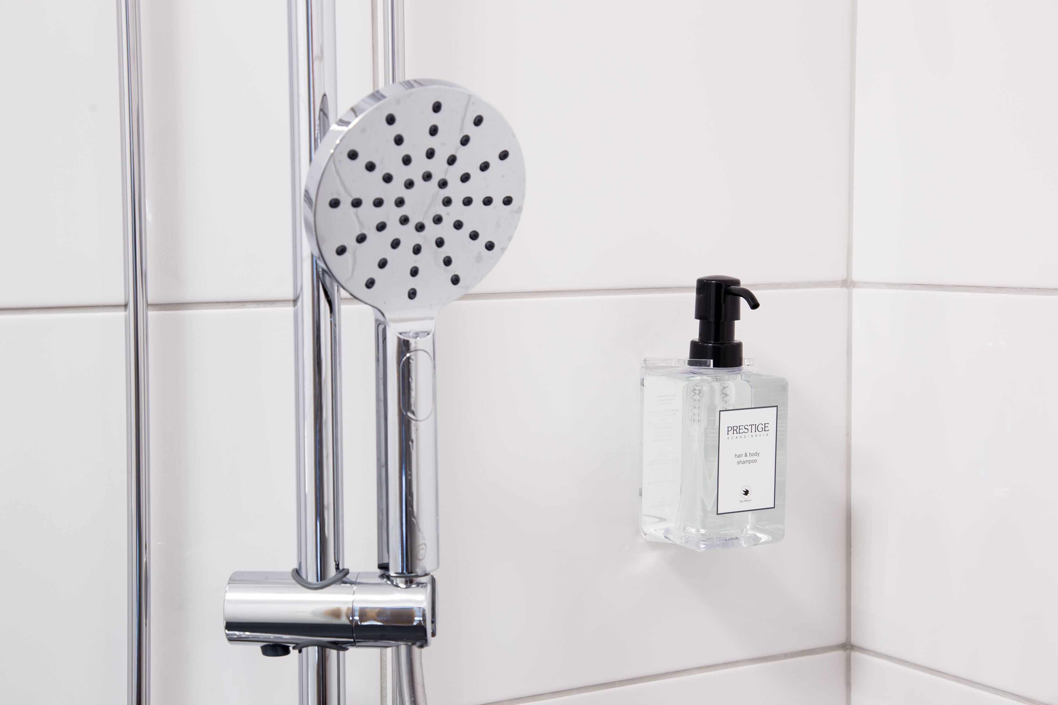 A chrome showerhead and a bottle of Prestige hair and body shampoo in a bathroom with white tiled walls.