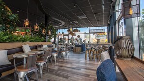 Interior moderno de restaurante con suelo de madera, sillas de metal y mesas. Hay luces colgantes y un jarrón grande sobre una mesa de madera.