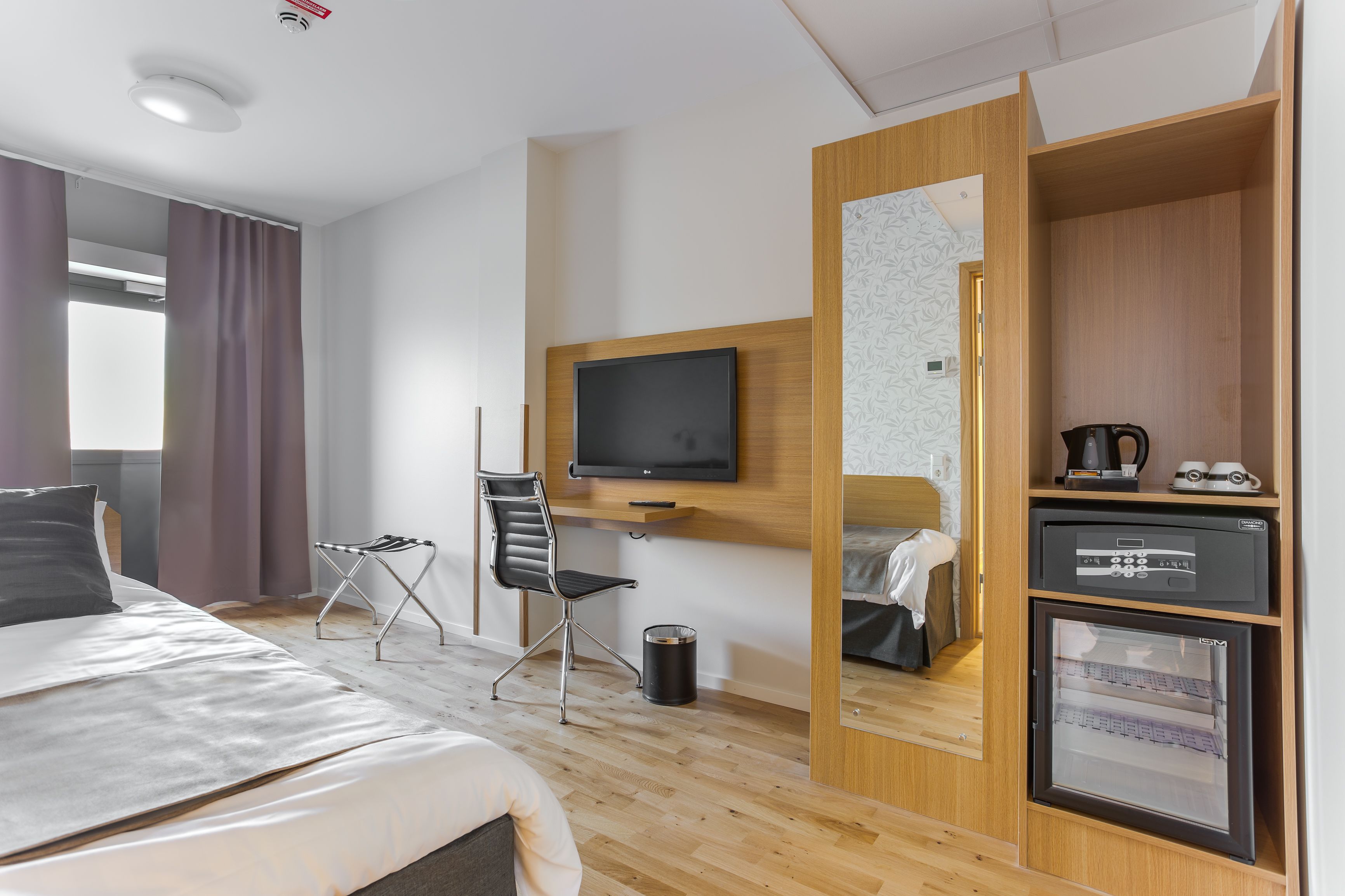 A hotel room with a bed, a TV mounted on the wall, a desk with a chair, a mini-fridge, and a trash can.