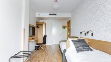 Ein Hotelzimmer mit Holzboden, einem Bett mit weißer und grauer Bettwäsche, einem Sessel, einem Fernseher auf einer Holzablage und einem Wandpapier mit Muster.
