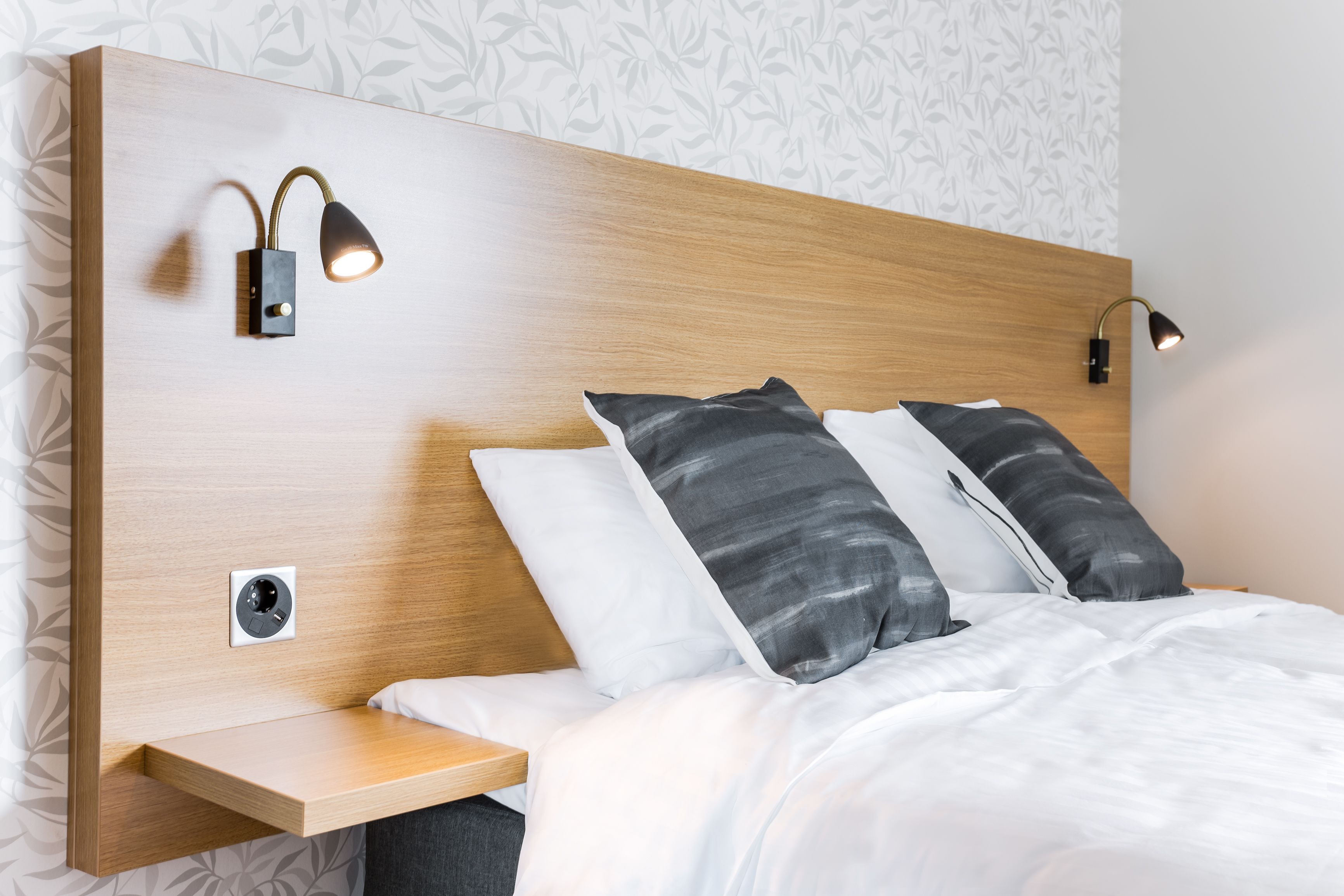 A modern bedroom with a wooden headboard, two bedside lamps, a nightstand, and a bed with white and gray bedding.