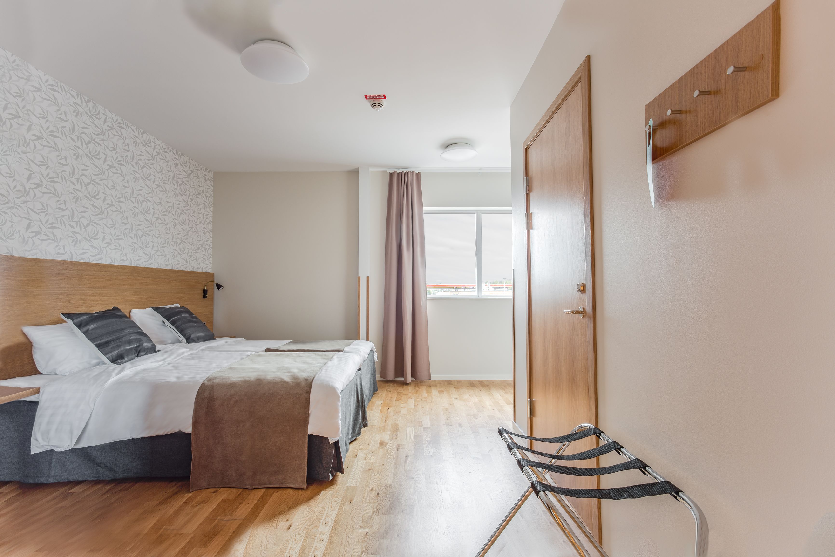 A hotel room with a wooden floor, a bed with a white and gray duvet, a wooden headboard, a window with curtains, and a coat rack.