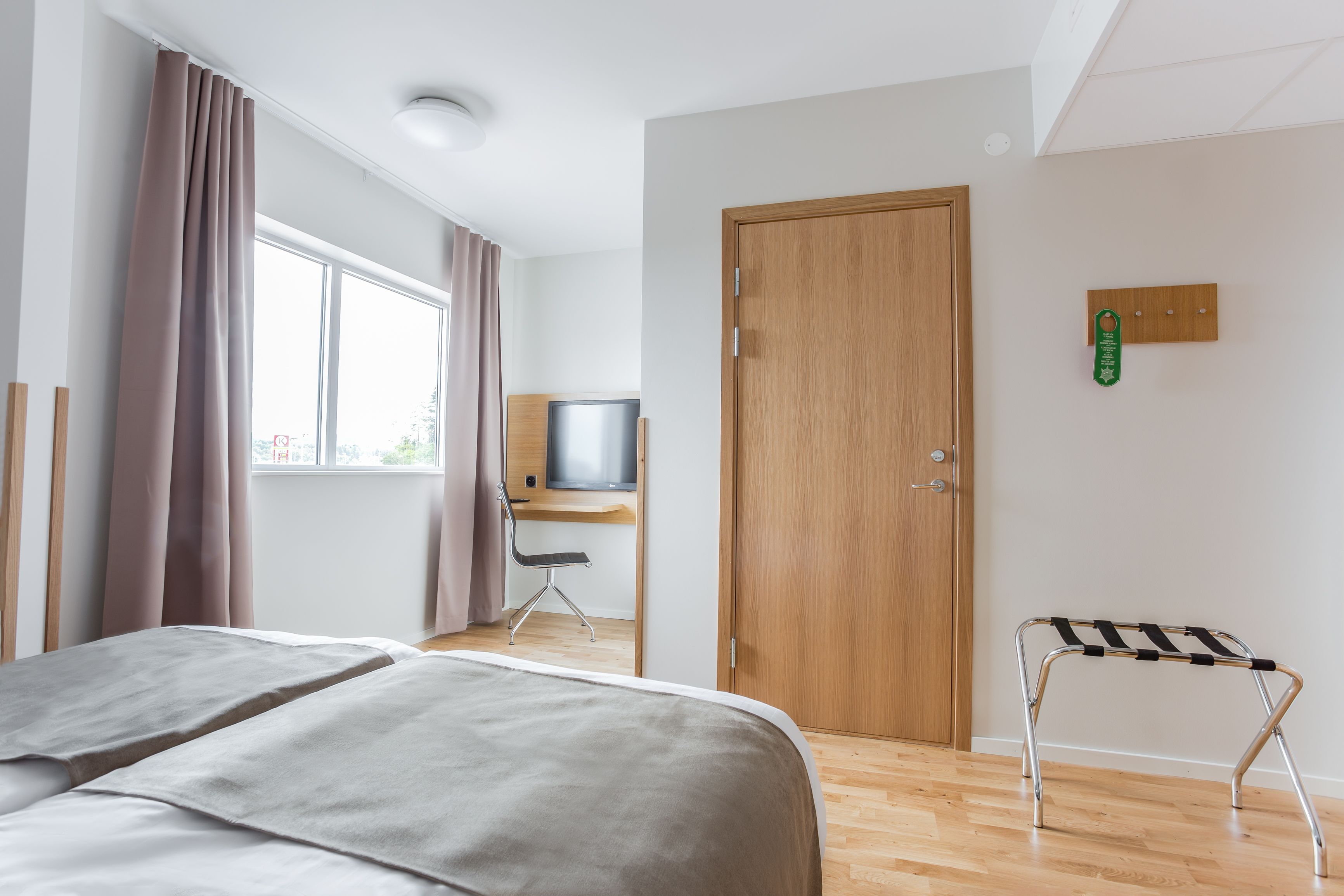 Quarto de hotel com cama, escrivaninha e cadeira, televisão, porta de madeira e pequena mesa com tecido listrado em preto e branco.