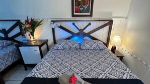 A hotel room with two beds, a headboard with a blue light, a framed painting on the wall, and a bedside table with a lamp. - Hotel Maritza (Cahuita)