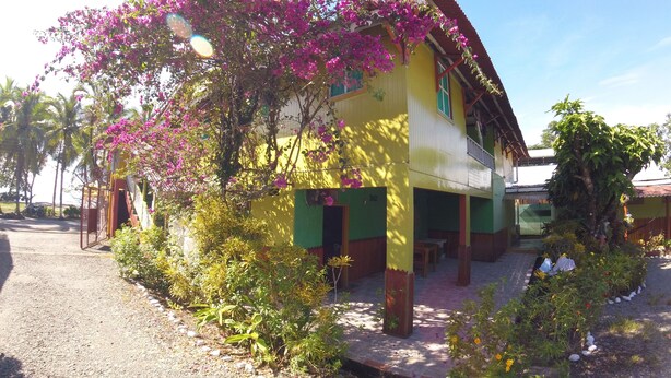 A two-story building with a yellow facade and green trim, surrounded by a gravel driveway and various plants. - Hotel Maritza (Cahuita)