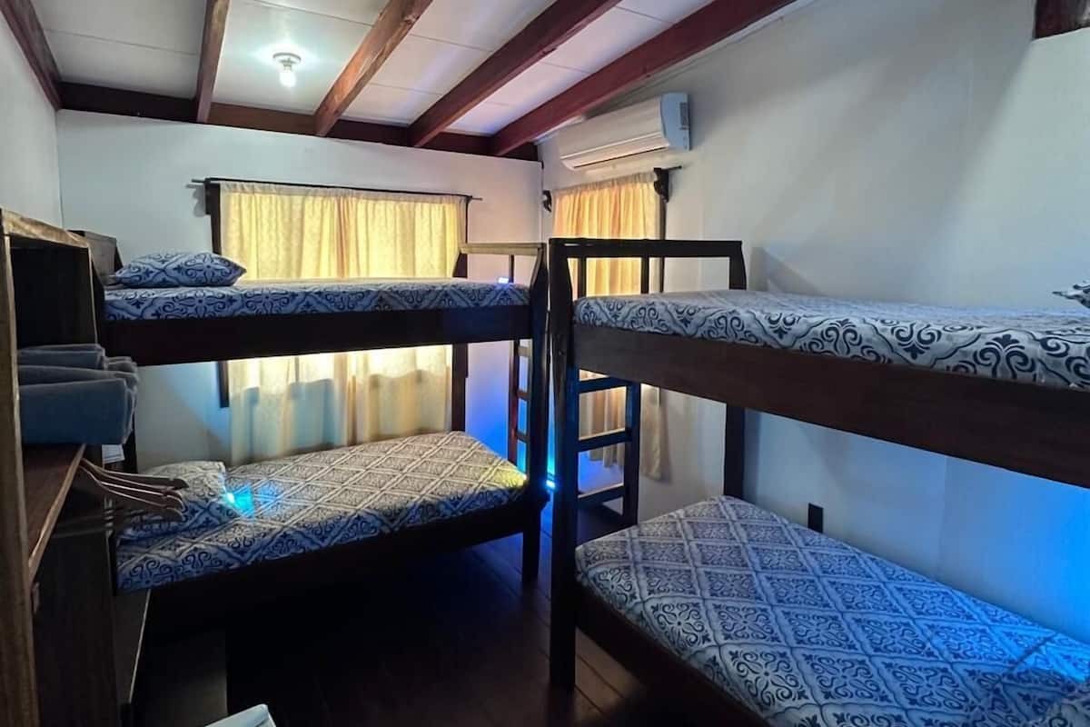 A dormitory room with bunk beds, wooden beams, a ceiling light, and an air conditioning unit.