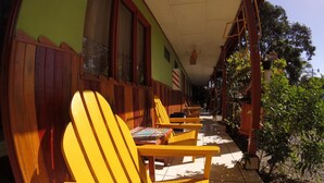 A covered outdoor seating area with wooden chairs and tables, a tiled floor, and a green wall with windows. - Hotel Maritza (Cahuita)