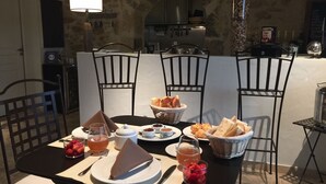 A dining table set with plates, glasses, and a variety of food items, surrounded by chairs in a room with stone walls and a kitchen in the background. - La Rivayne (Lauris)