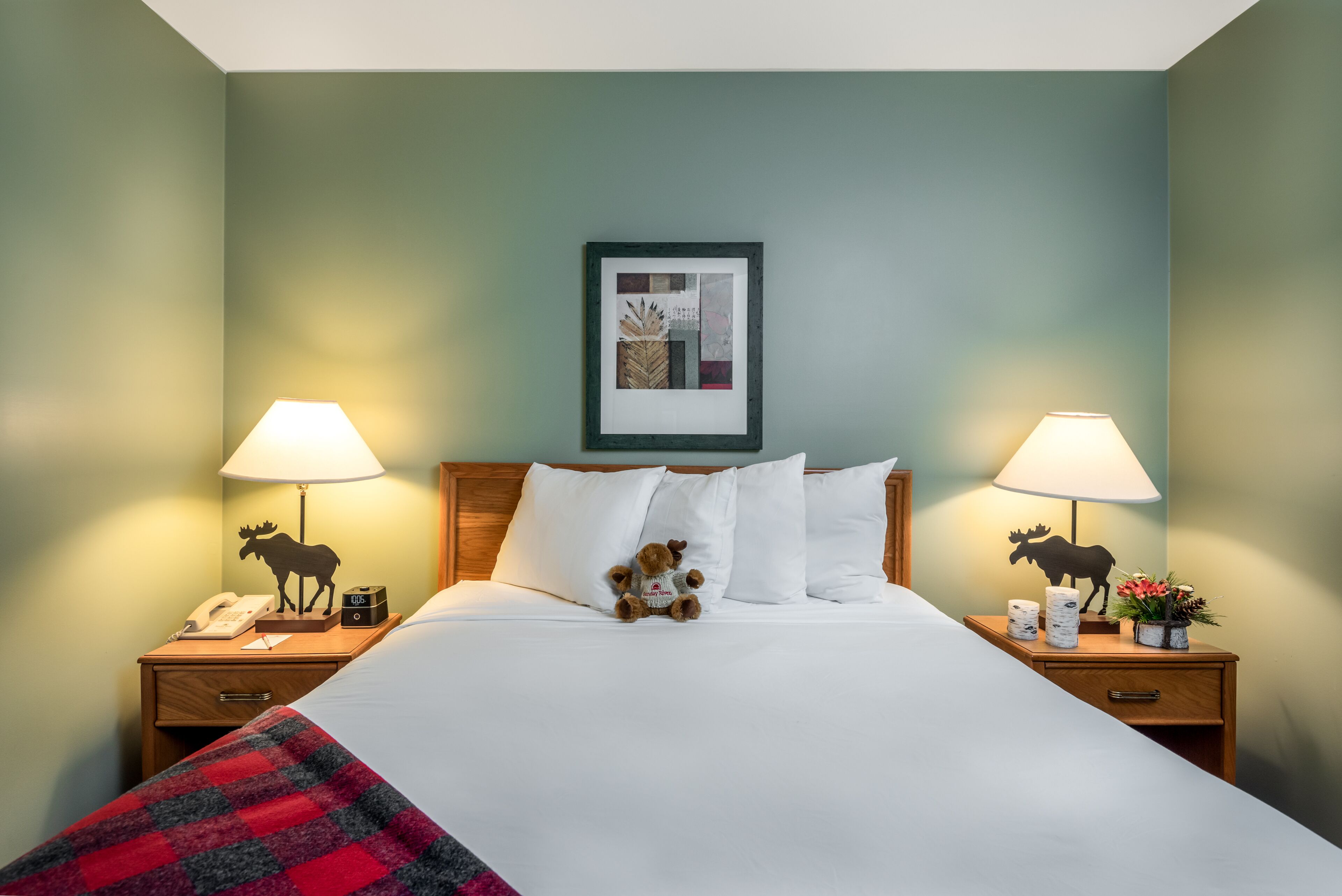 A neatly made bed with white linens, flanked by two bedside tables with lamps, each table featuring a moose silhouette. A framed artwork hangs above the bed, and a red and black checkered throw is draped over the foot of the bed.