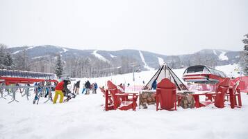 Área externa coberta de neve, com cadeiras e mesas vermelhas, um teleférico e montanhas ao fundo.