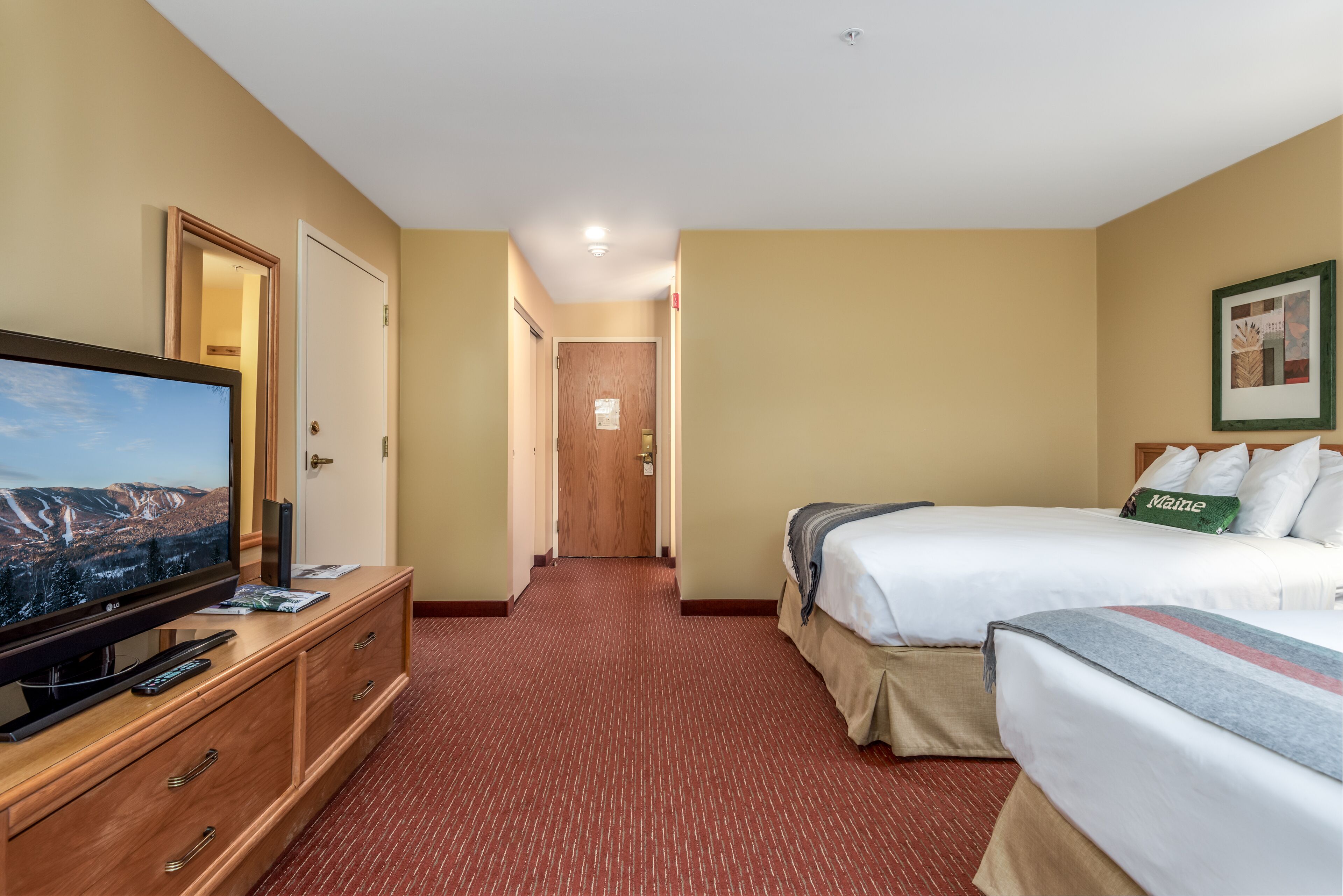 A hotel room with a television on a wooden dresser, a bed with a 'Maine' pillow, and a framed picture on the wall.