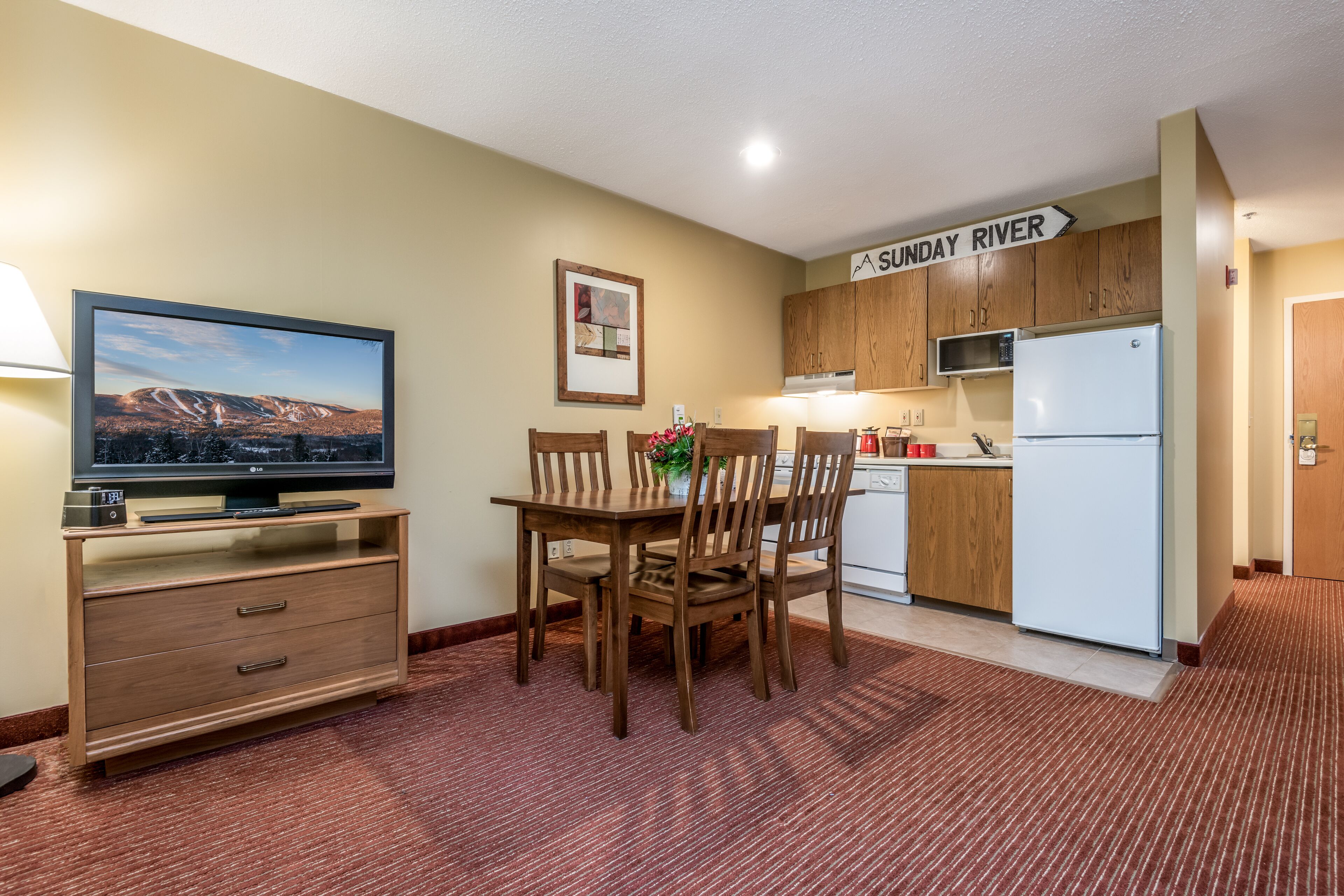 A compact kitchen with wooden cabinets, a microwave, and a refrigerator. There's a dining table with chairs and a flat-screen TV on a wooden stand. A sign on the wall reads "SUNDAY RIVER."