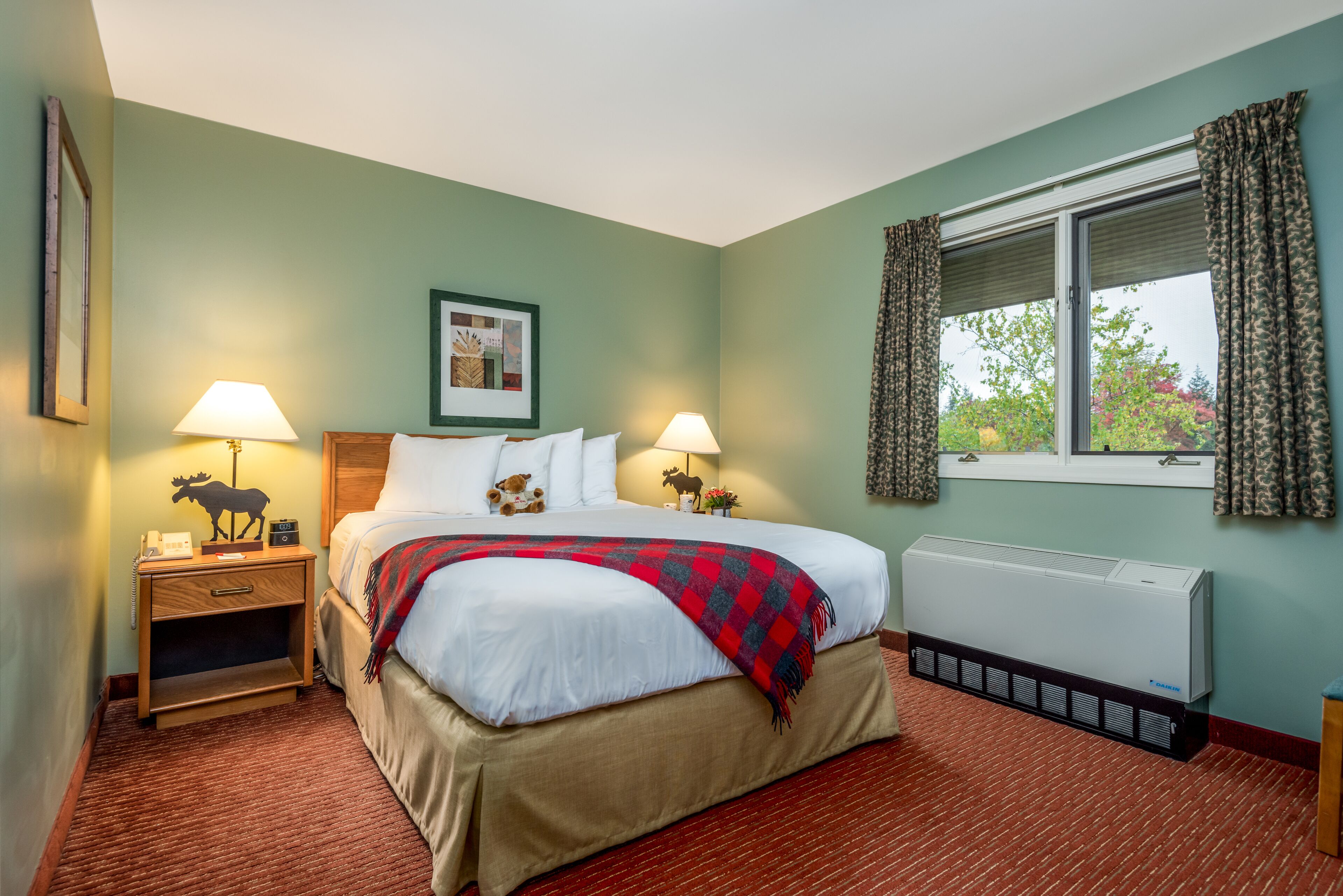 Una habitación de hotel con una cama, dos mesitas de noche con lámparas, un cuadro en la pared, una ventana con cortinas y un radiador.