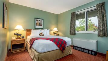 Una habitación de hotel con una cama, dos mesitas de noche con lámparas, un cuadro en la pared, una ventana con cortinas y un radiador.